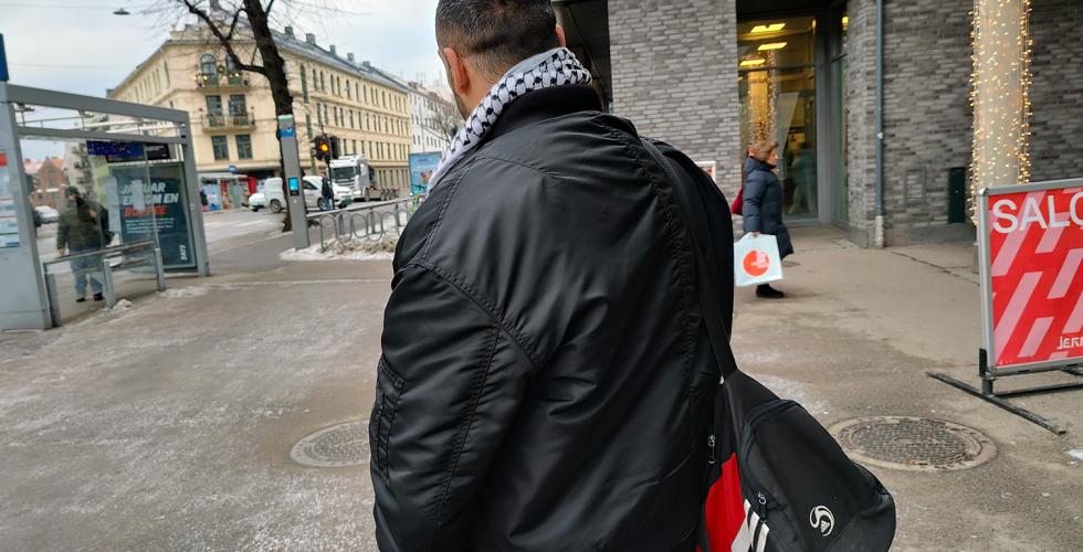 Mann med ryggen til. Sort jakke. Sort bag. Gr&aring;tt rundt.