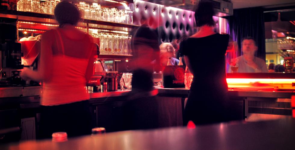 Rosa lys i en bar. Noen bartendere st&aring;r og shaker drinker, mens kundene venter. 