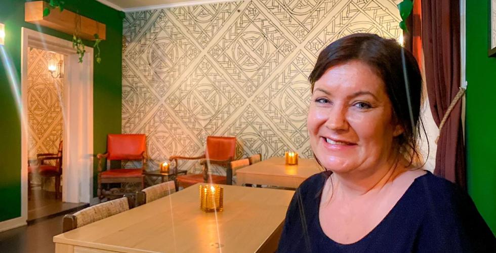 Ei smilende dame st&aring;r p&aring; en sofa og har p&aring; seg en svart t-skjorte. 