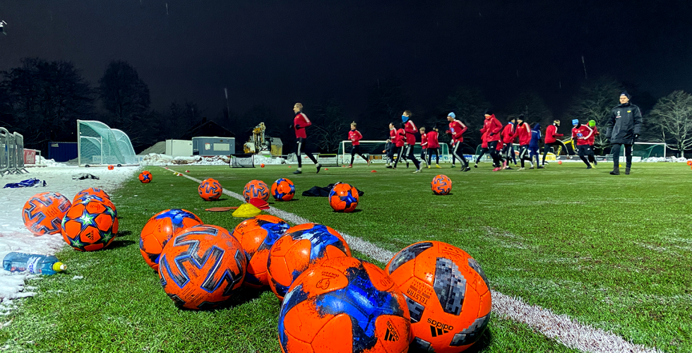 P&aring; fotballbane med spillere som trener i bakgrunn. Ser fotballer og en drikkeflaske.