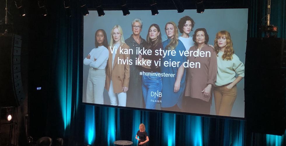 Aina Lemoen Lunde foran et stor publikum p&aring; Sentrum Scene i Oslo, med DNBs reklame for #HunInvesterer p&aring; lerret bak seg.