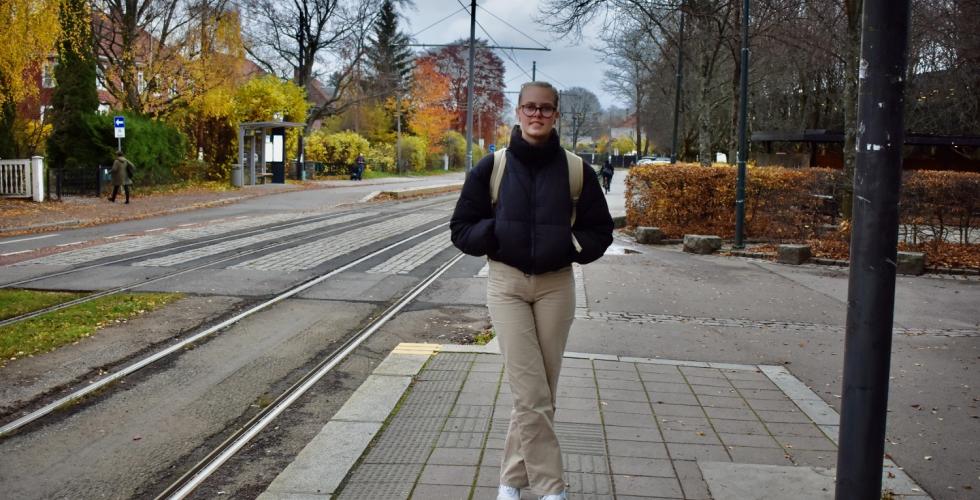 Blond kvinne med svart jakke, beige ryggsekk og m&oslash;rke briller st&aring;r og venter p&aring; trikken utenfor Universitetet i Oslo, avd. Blindern