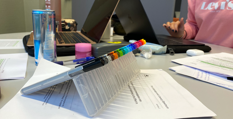 Bilde av et rotete bord med to pcer, b&oslash;ker, tusjer og energidrikk. To jenter som sitter og jobber med eksamen