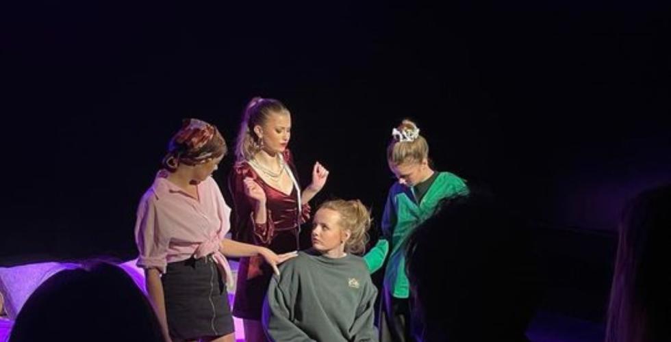 P&aring; en liten teaterscene st&aring;r ei jente i posete kl&aelig;r. Rundt henne st&aring;r fullt sminkede jenter i trange kl&aelig;r og trykker seg p&aring; henne. 