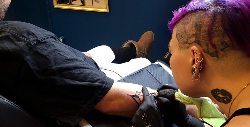 Ei dame tatoverar eit lyn p&aring; armen til ein mann. Tatov&oslash;ren har lilla h&aring;r.