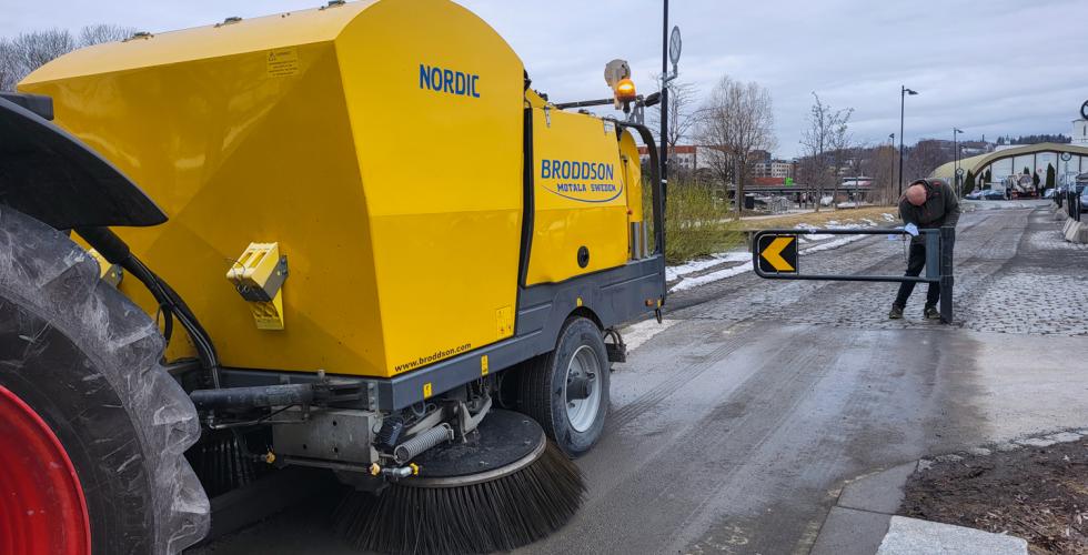 Bilde av hengeren p&aring; en feiebil, som er stor og gul. I bakgrunnen er det en mann som lukker en bom.