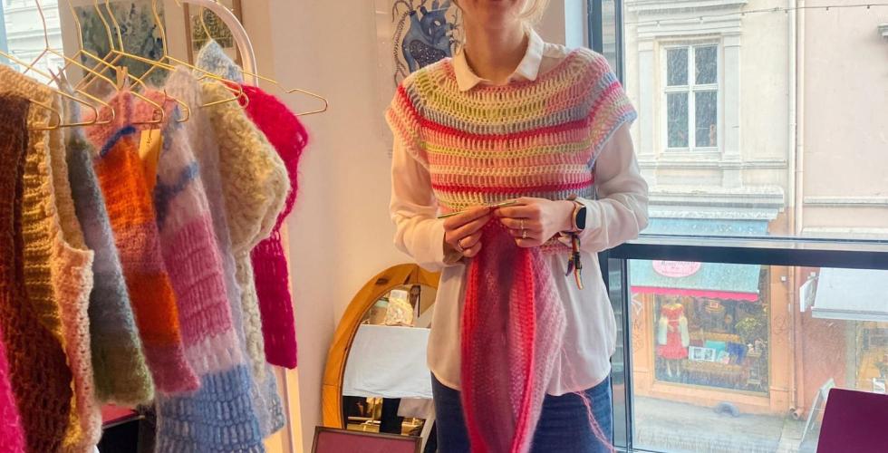 Ei jente med briller foran et stand med fargerike kl&aelig;r. Hun har p&aring; seg en hvit skjorte, og over en fargerik vest. 