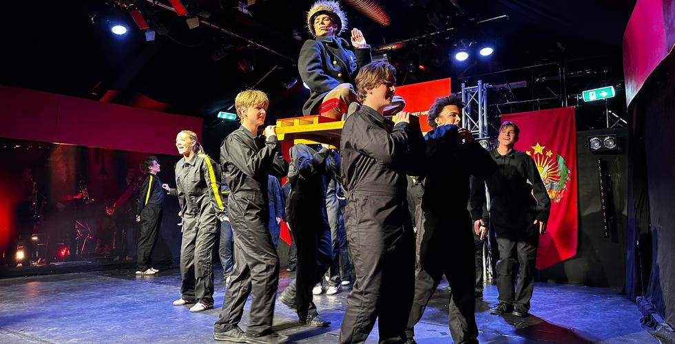 Akt&oslash;rer i svarte drakter g&aring;r p&aring; en scene og b&aelig;rer en annen akt&oslash;r p&aring; en b&aring;re. 