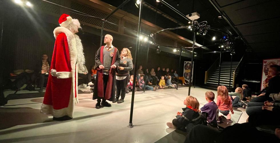 To skuespillere utkledt som Harry Potter og Hermine i et lite lyssatt scenerom. De ser p&aring; en mann i julenissedrakt. I enden av rommet er en hvitkledd trapp. I taket er det lys. Langs sidene sitter det barn p&aring; matter. 