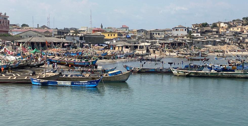 vi ser et bl&aring;tt vann med b&aring;ter, en strand og flere hus i bakgrunnen