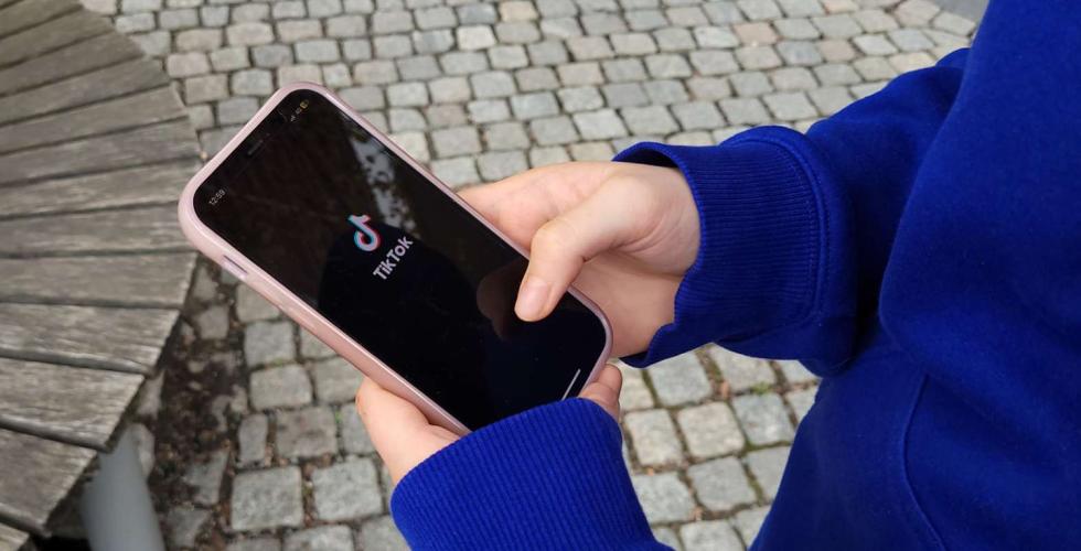 Bildet viser en jente som ser p&aring; telefonen sin. Hun har TikTok oppe og logoen til TikTok vises p&aring; skjermen. Jenta har p&aring; seg bl&aring; genser og st&aring;r ute. 
