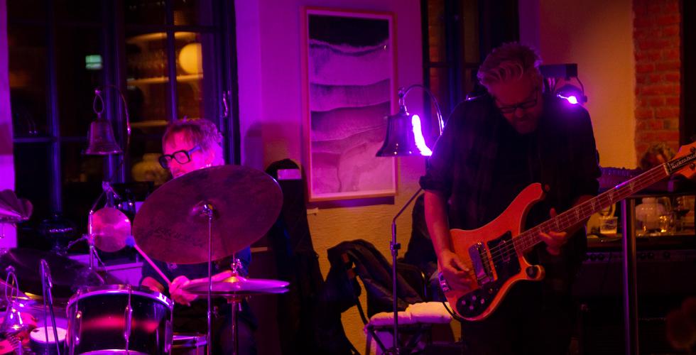 En mann med svarte briller sitter bak et trommesett. En mann med gr&aring;tt h&aring;r spiller bass. 