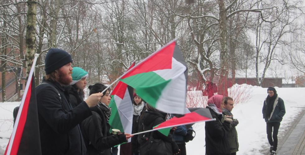 Flere personer er i bildet, p&aring; en uformell rekke. De holder palestinske flagg, og ser konsentrert til rett frem mot taleren. 