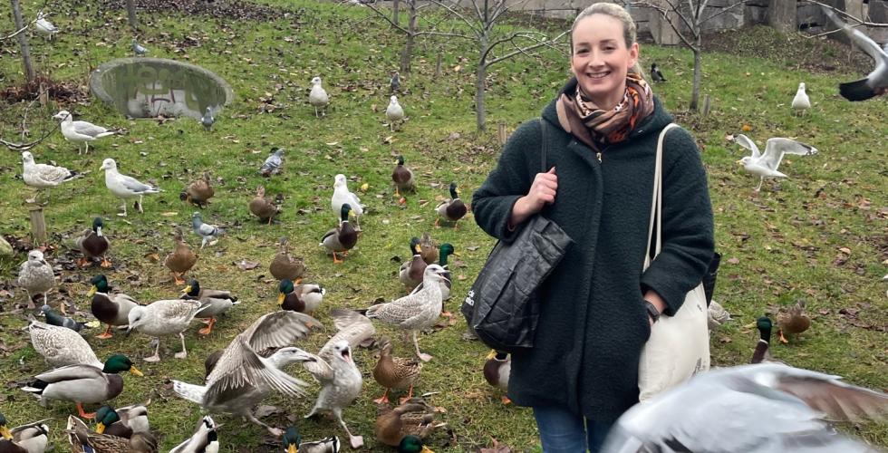 Mai Amundsen st&aring;r og smiler til kameraet. Rundt henne g&aring;r det en haug med ulike fugler, blant annet m&aring;ker og ender.