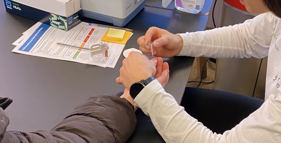 Bildet viser en kvinne som f&aring;r sjekket blodsukkeret sitt. Hun har blitt stukket i fingeren, s&aring; det er litt blod p&aring; fingeren. Frisklivsveilederen holder et utstyr til &aring; hente noen dr&aring;per til m&aring;lingen.