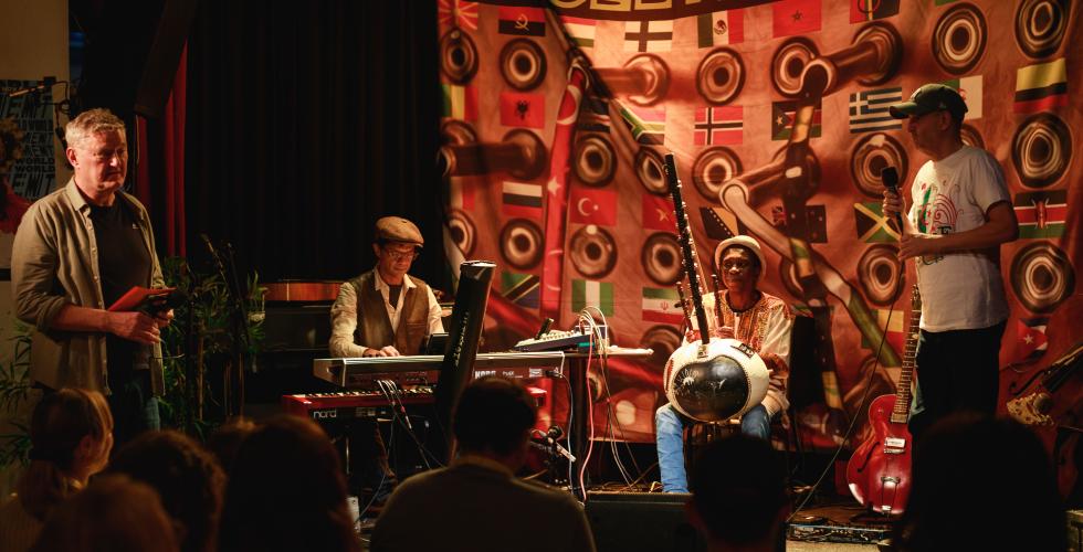 Fire personer p&aring; en scene, de to i midten sitter med hvert sitt instrument, og de to p&aring; sidene st&aring;r. Han ene - Arne Berg, programleder for Jungeltelegrafen - st&aring;r med en mikrofon og noen lapper i h&aring;nda, han andre som st&aring;r har og mikrofon.