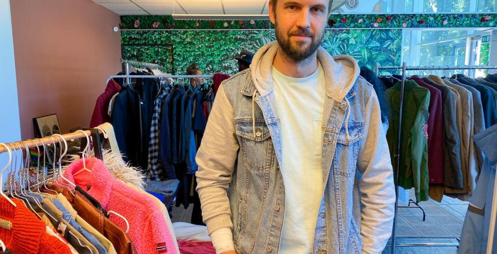 Mannen st&aring;r foran noen kl&aelig;rstativer med hendene i lomma. Han har p&aring; seg en hvit tskjorte med en denim jeansjakke over.