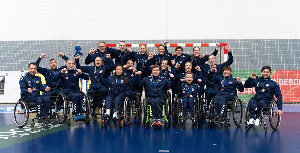 Alle spillerne p&aring; rullestolh&aring;ndballandslaget og st&oslash;tteapparatet bak st&aring;r foran et m&aring;l og jubler med bronsemedaljen rundt halsen