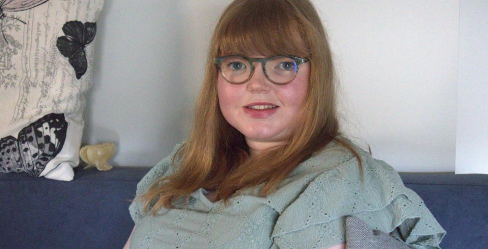 Kvinne p&aring; 31 &aring;r med r&oslash;dt h&aring;r og briller sitter i gr&aring; sofa og ser p&aring; kameraet.