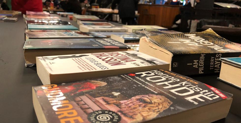 mange b&oslash;ker ligger p&aring; et bord. Personer st&aring;r i bakgrunnen