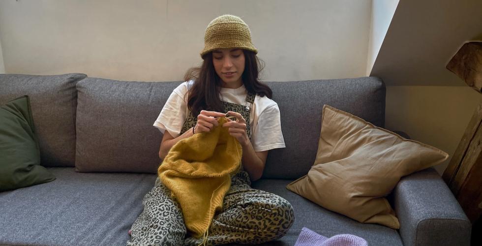 Nesrin Alma Kalayci sitter i sofaen og strikker p&aring; en gul genser. 