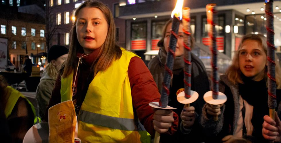 Jente ser i kamera mens hun holder en opptent fakkel. Hun tenner faklene til to andre ogs&aring;.