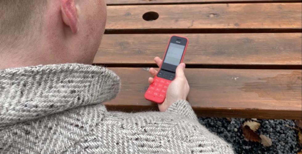 Bilde av en mann med gr&aring; ullk&aring;pe som sitter p&aring; en benk. Han holder en r&oslash;d Nokia - knappetelefon. 