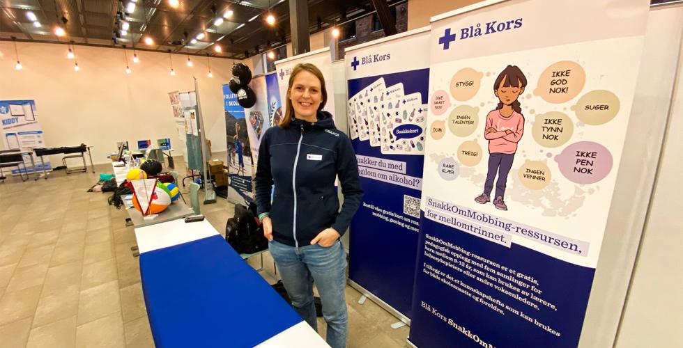 En kvinne i bl&aring; genser st&aring;r foran et bord. 