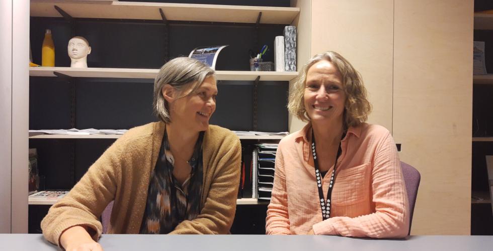 To kvinner som sitter. Den ene ser p&aring; den andre, mens den andre ser i kamera.