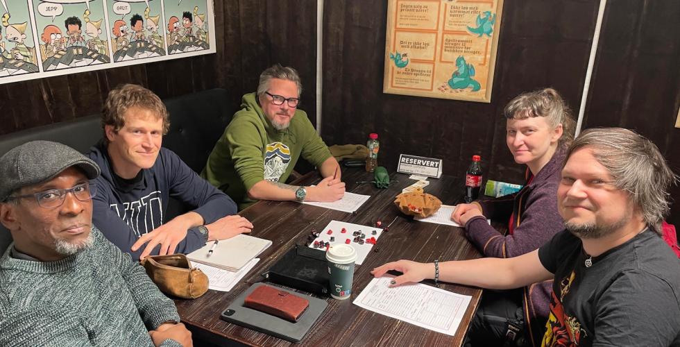 Fem mennesker som sitter rundt et bord og spiller rollespillet Vampire the Masquerade. P&aring; bordet ligger det terninger og ark med rollebeskrivelser.