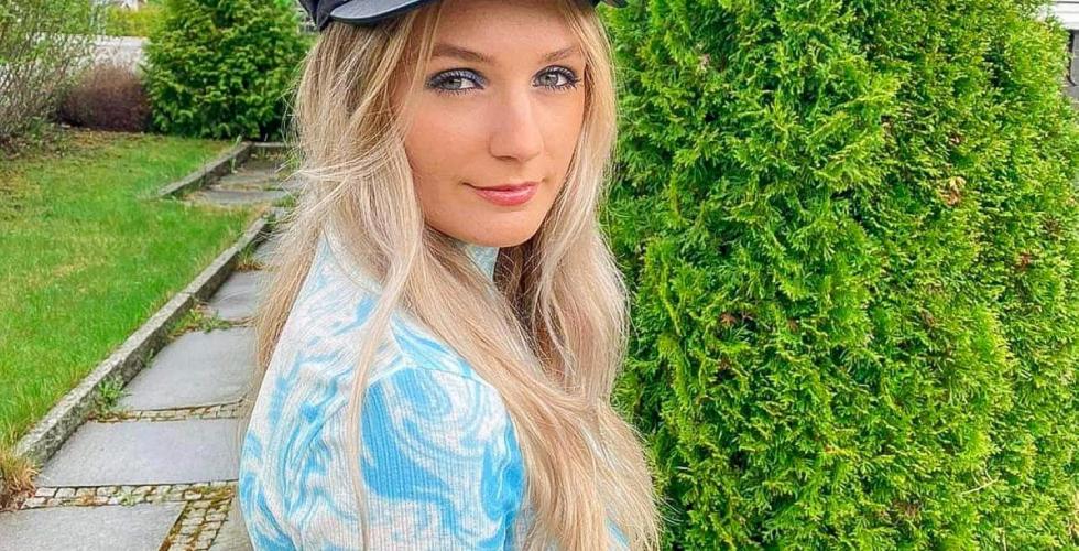 Sanna har p&aring; seg en svart lue og st&aring;r foran noen busker. Hun har p&aring; seg en lysebl&aring; genser med hvite b&oslash;lger. Hun har langt, blondt h&aring;r og smiler til kamera. 