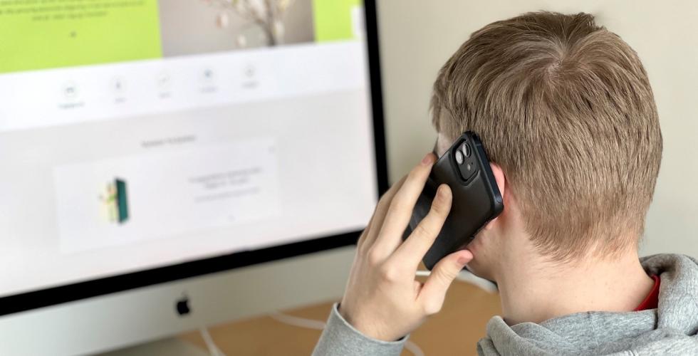 Person sitter med telefonen mot &oslash;re og ser p&aring; PCen. 