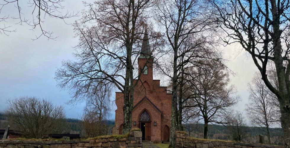 Bildet viser S&oslash;rkedalen kirke i Oslo. Kirken er laget av brune murstein, og har et h&oslash;yt kirkespir. Den st&aring;r litt tildekket av nakne l&oslash;vtr&aelig;r. 