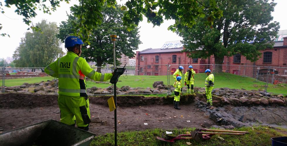 En gruppe mennesker st&aring;r i bakgrunnen med verneutstyr og snakker. En dame st&aring;r i forgrunnen med en type m&aring;leinstrument og har ogs&aring; p&aring; seg verneutstyr.